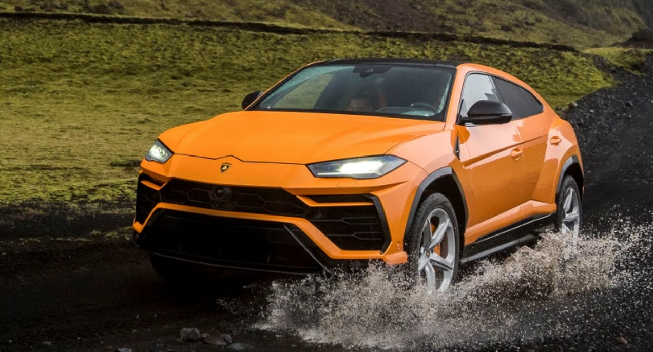 An orange Lamborghini Urus SUV is driving through shallow water.