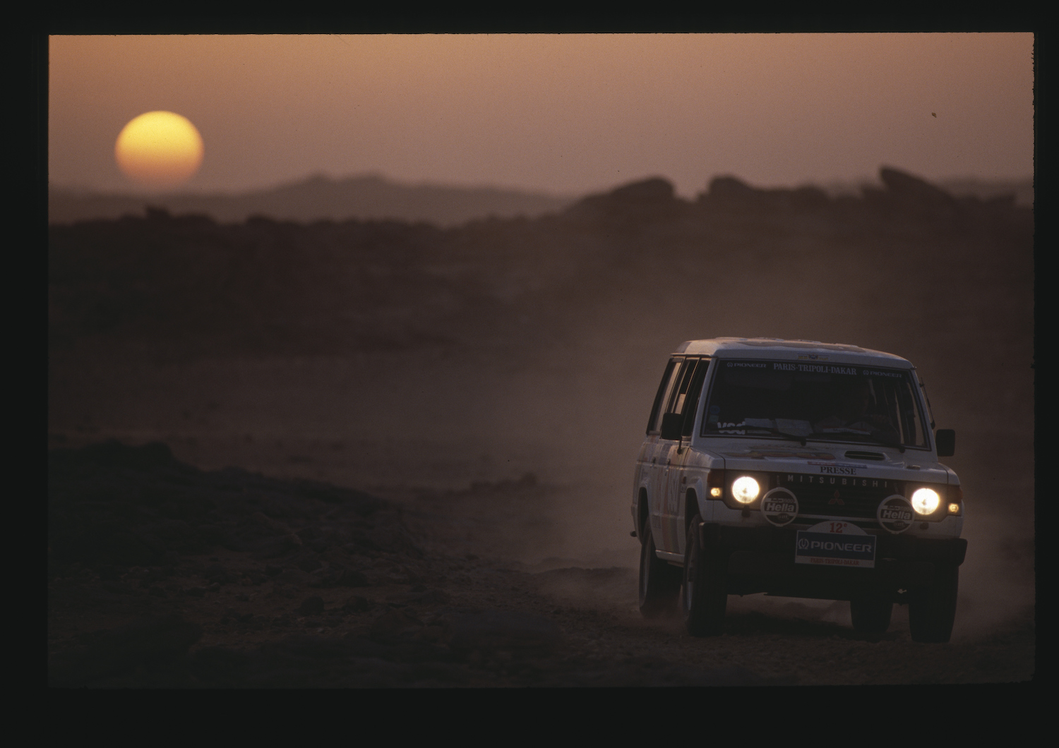 Mitsubishi Montero SUV racing through the desert on an early Paris-Dakar rally, the sun setting behind a ridge of mountains visible behind it.