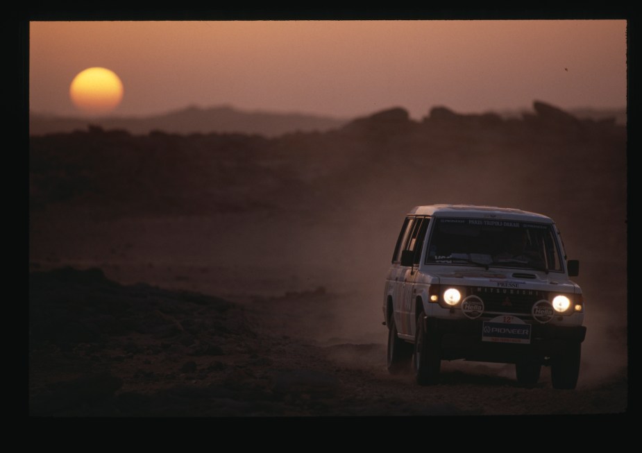 Mitsubishi Montero SUV racing through the desert on an early Paris-Dakar rally, the sun setting behind a ridge of mountains visible behind it.