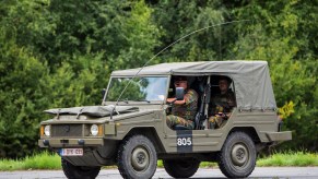 Soldier driving a military-spec Volkswagen type 183 Iltis on pavement, a wall of trees visible in the background.