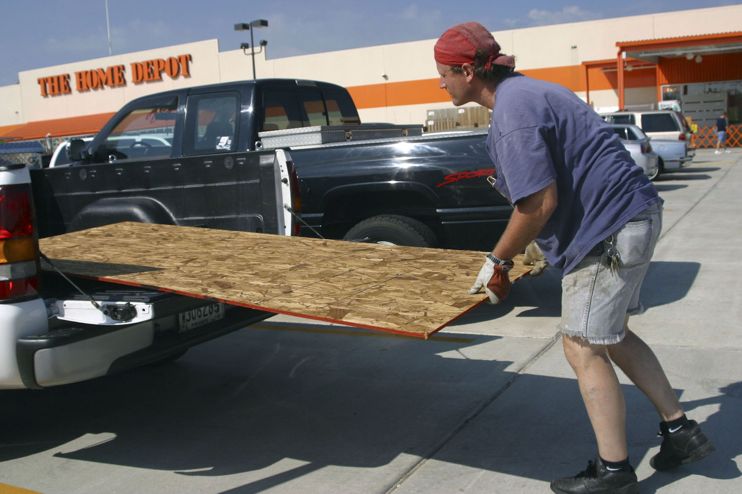 A man slides a sheet of plywood into the bed of a pickup truck, Home Depot visible behind him.