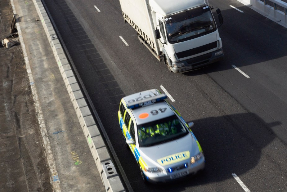A police car passes a lorry on a UK motorway, the road visible behind.