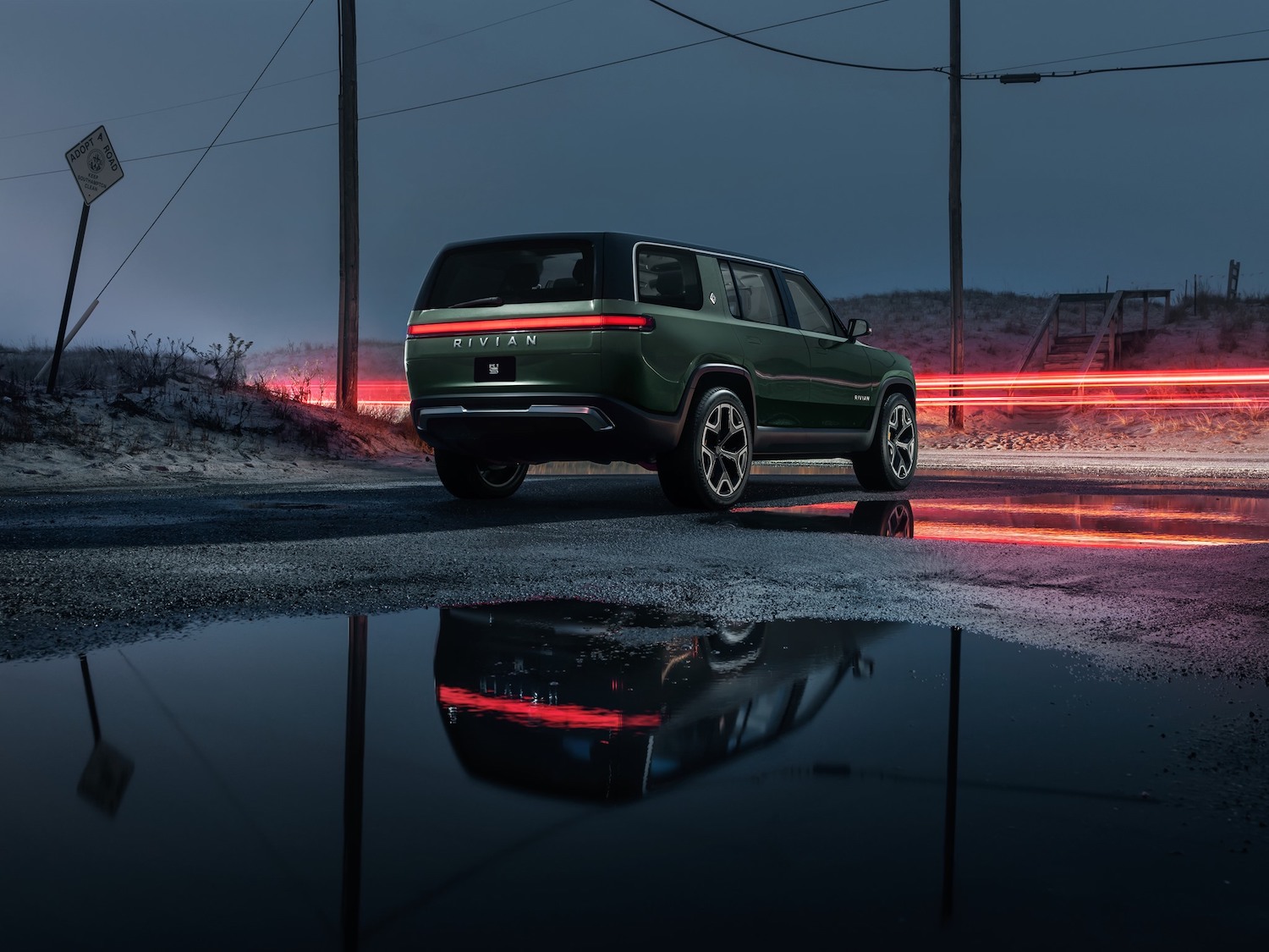 Promo photo of a green Rivian SUV EV parked next to a road, tail lights visible in the background.