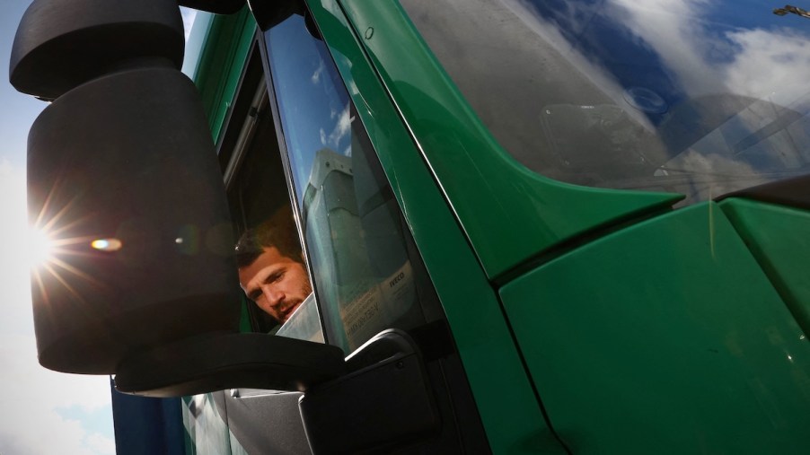 A truck driver, potentially with truck driver sun damage, in a green truck with a sunspot on the mirror.