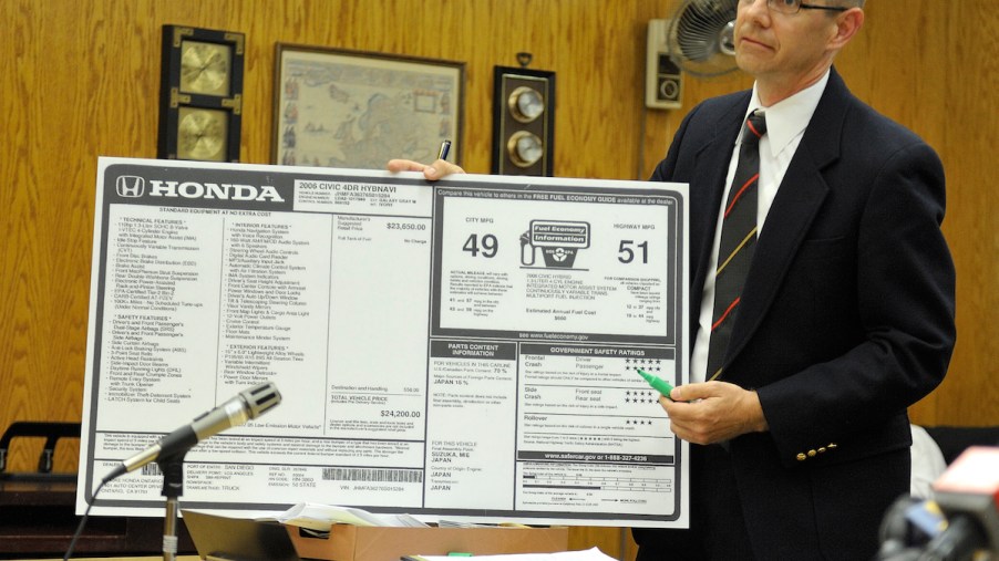 A Dealership sticker that displays the information regarding the car, including the fuel economy ratings, being held by a man in a wood paneled office.