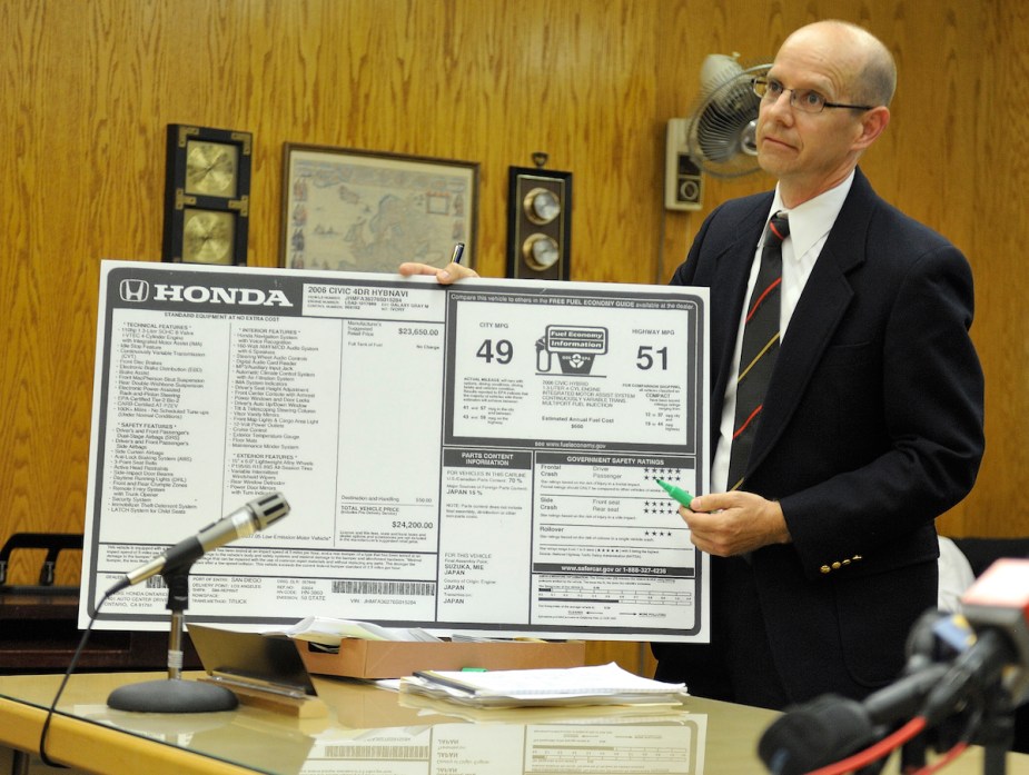 A Dealership sticker that displays the information regarding the car, including the fuel economy ratings, being held by a man in a wood paneled office. 