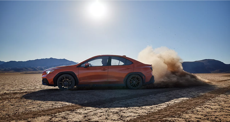 An orange 2022 Subaru WRX parked on a dry and cracked dirt plain under the sun and near a cloud of dust