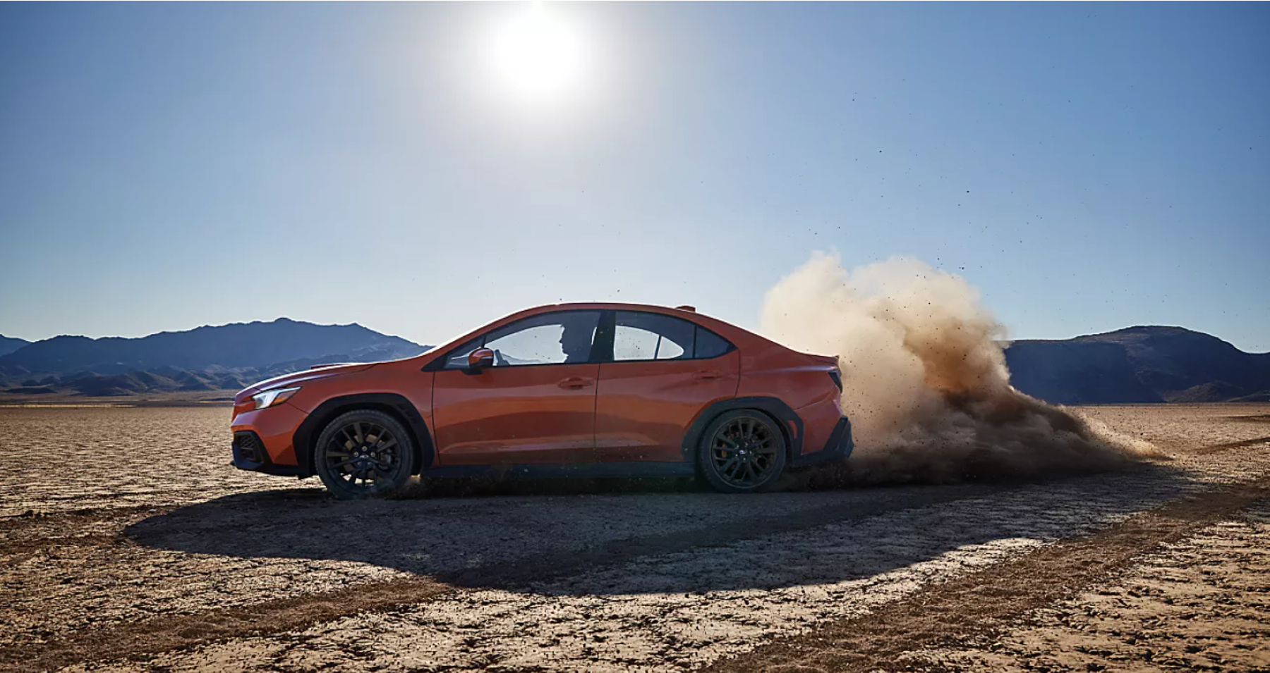 An orange 2022 Subaru WRX parked on a dry and cracked dirt plain under the sun and near a cloud of dust