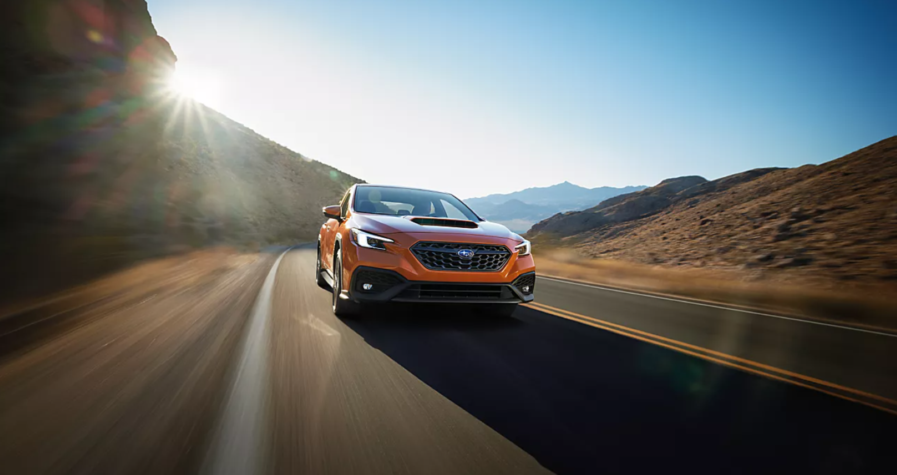 An orange 2022 Subaru WRX driving on a desert highway near rolling hills as the sun glares in a cloudless sky