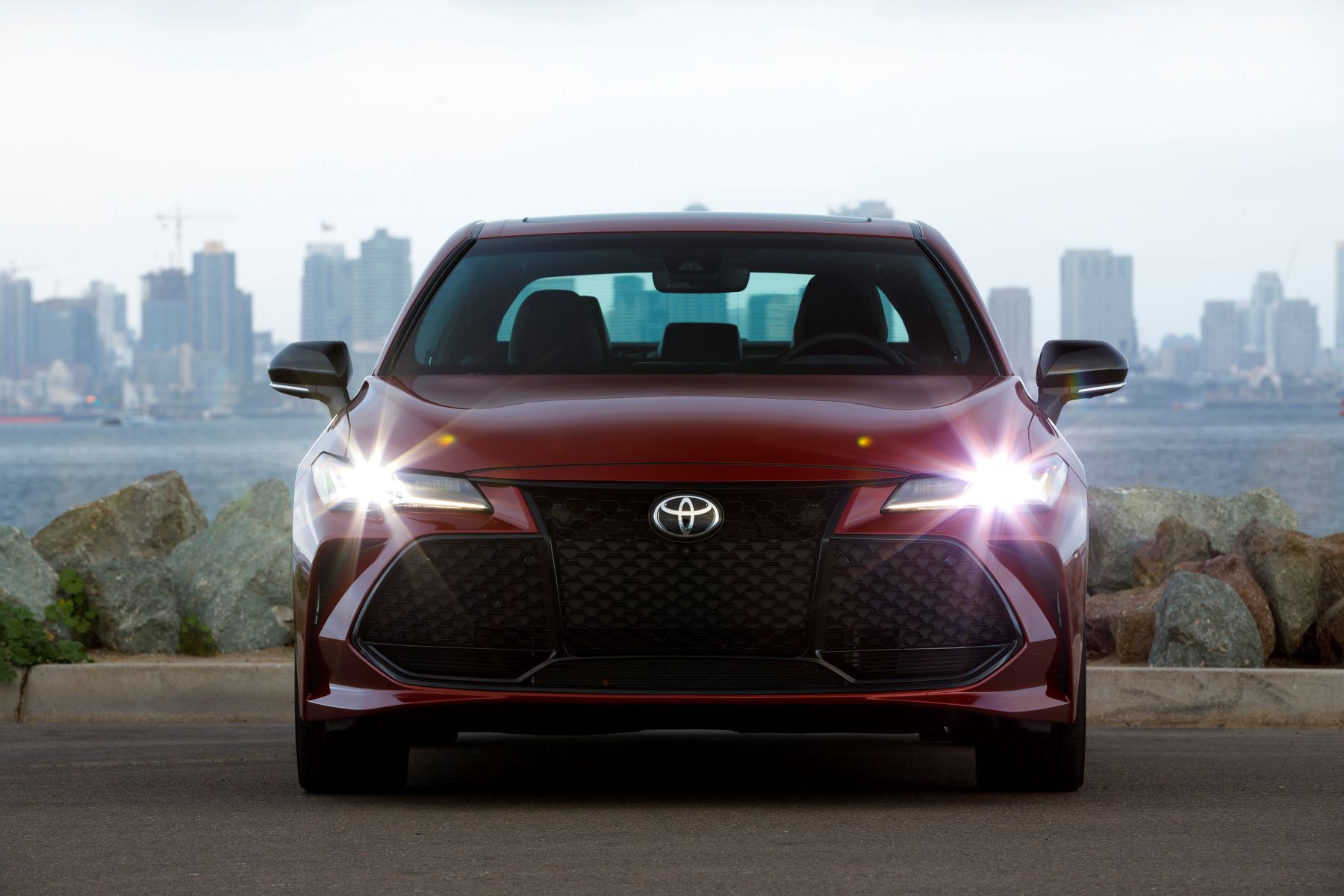 A red 2022 Toyota Avalon Touring full-size sedan model with its headlights on parked near a city skyline sea