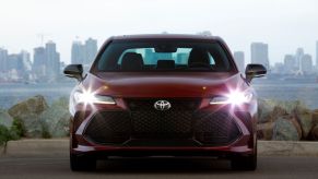 A red 2022 Toyota Avalon Touring full-size sedan model with its headlights on parked near a city skyline sea