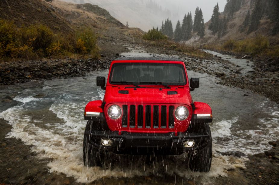 A red 2023 Jeep Wrangler off-road SUV driving through a muddy creek as it rains