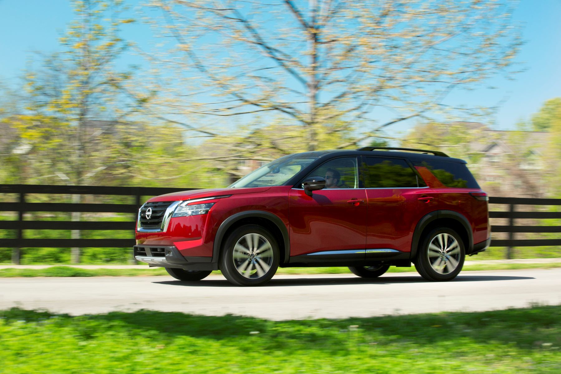 A red 2023 Nissan Pathfinder midsize SUV model driving down a country highway framed by wooden fencing