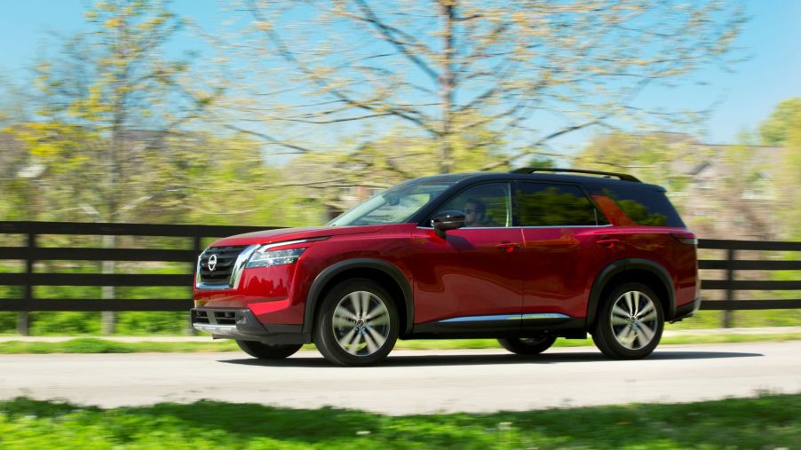 A red 2023 Nissan Pathfinder midsize SUV model driving down a country highway framed by wooden fencing