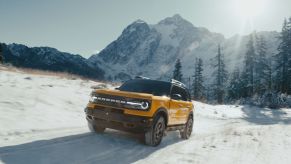 A yellow-gold 2022 Ford Bronco Sport driving off-road through a snowy field near a forest and mountains