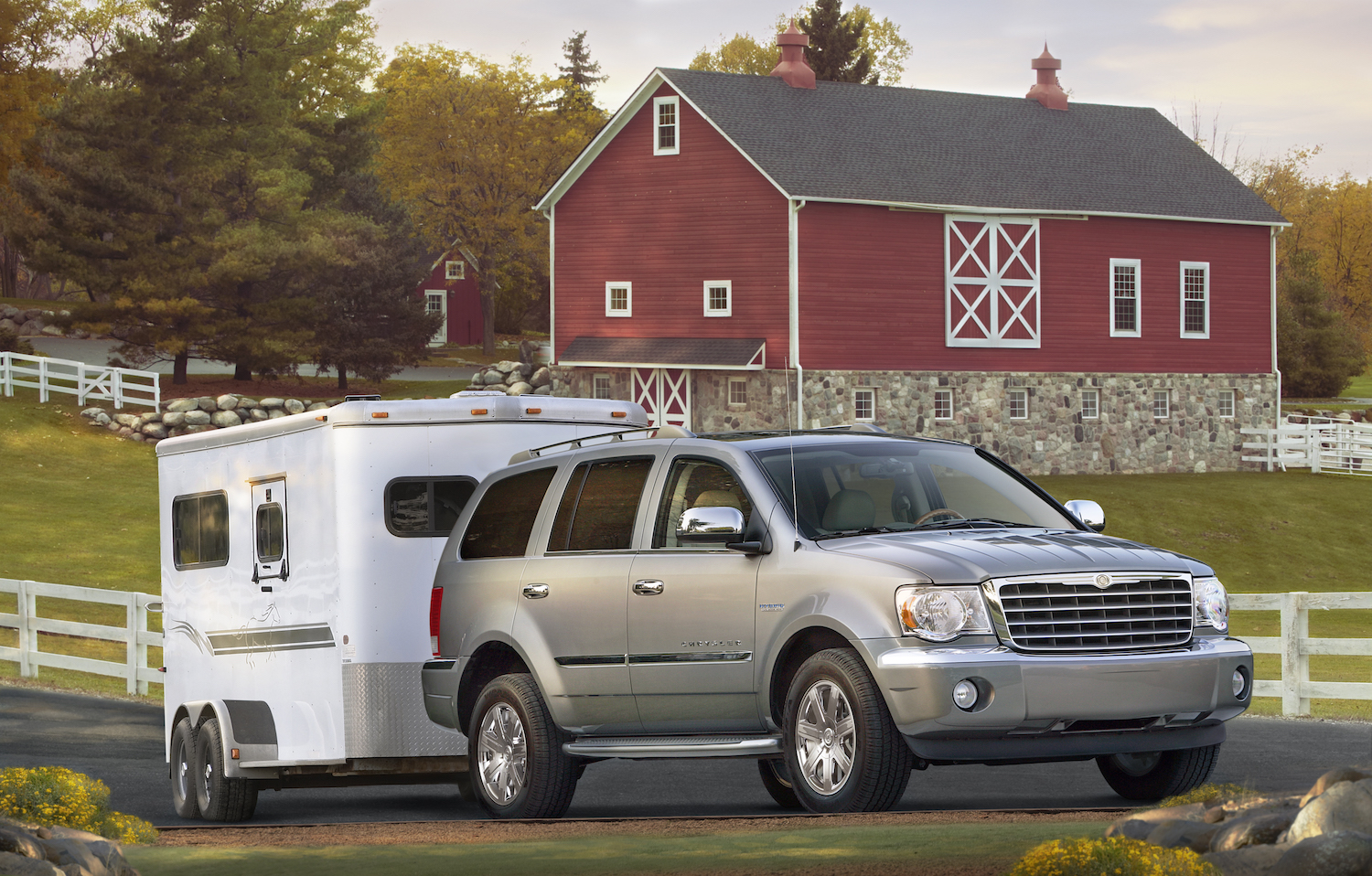 A 2009 Chrysler Aspen luxury full-size HEMI V8 hybrid SUV shows off its towing capacity, hitched to a trailer with a barn visible in the background.