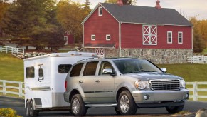 A 2009 hybrid Chrysler Aspen luxury full-size HEMI V8 powered SUV shows off its tow rating, hooked up to a trailer with a barn visible in teh background.