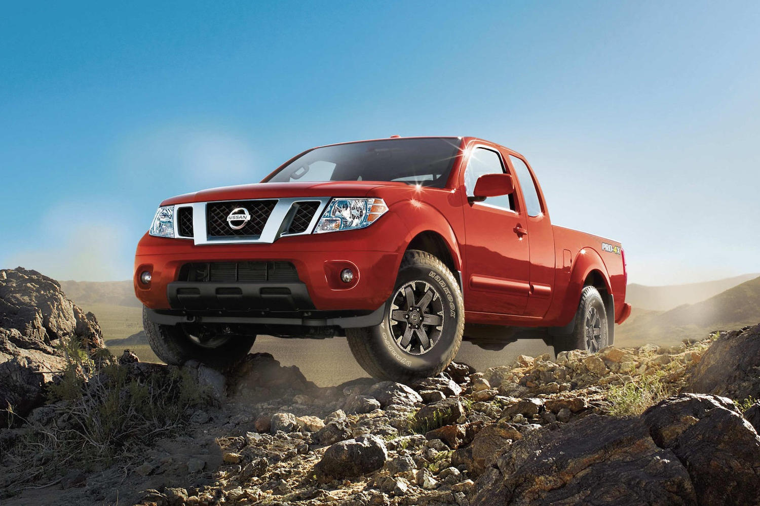 Promo photo of a red Nissan Frontier 4x4 reliable midsize pickup truck parked atop a rock pile, a cloudless blue sky visible in the background.