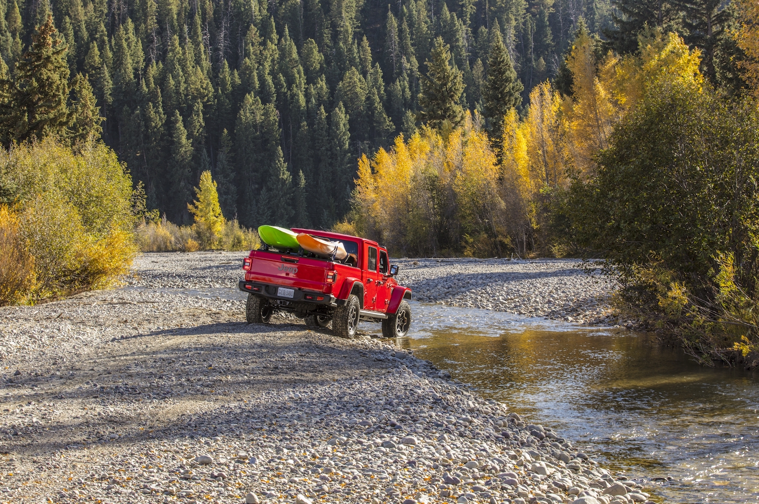 Red Jeep Gladiator pickup truck hauling kayaks through a river, trees visible in the background.