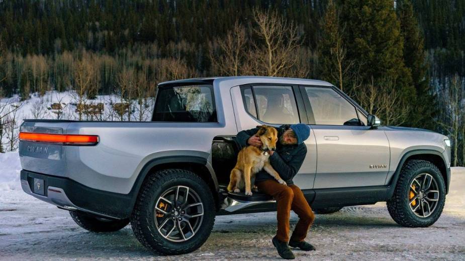 A man and a dog sit on the open door of a silver Rivian truck's gear tunnel storage compartment, a snowy hillside visible behind them.