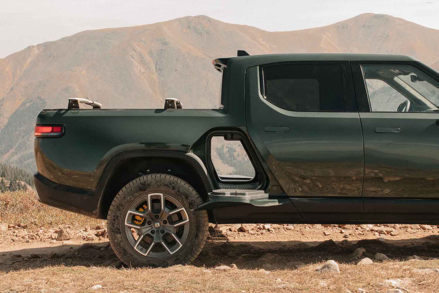 A Rivian electric truck parked in the desert with both its gear tunnel doors open so you can see all the way through the pickup, mountains visible in the background.