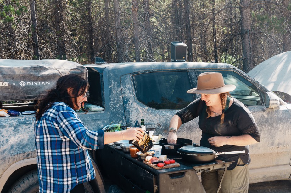 Two women lean over the unfolded camp kitchen built into the bed of a Rivian electric truck.