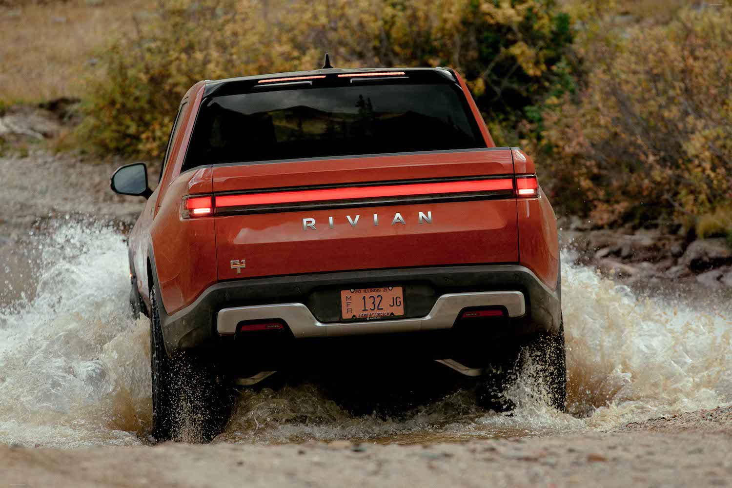 Red Rivian R1T electric pickup truck driving through a river, a field visible beyond it.