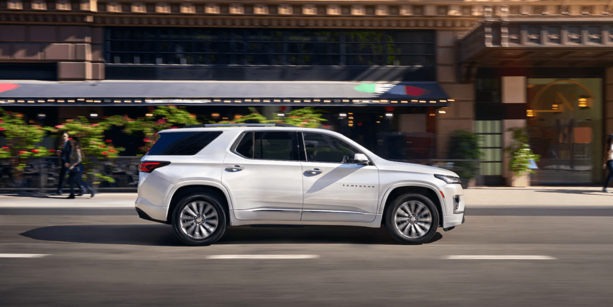 A side profile shot of a white 2023 Chevy Traverse full-size crossover SUV model driving through a city
