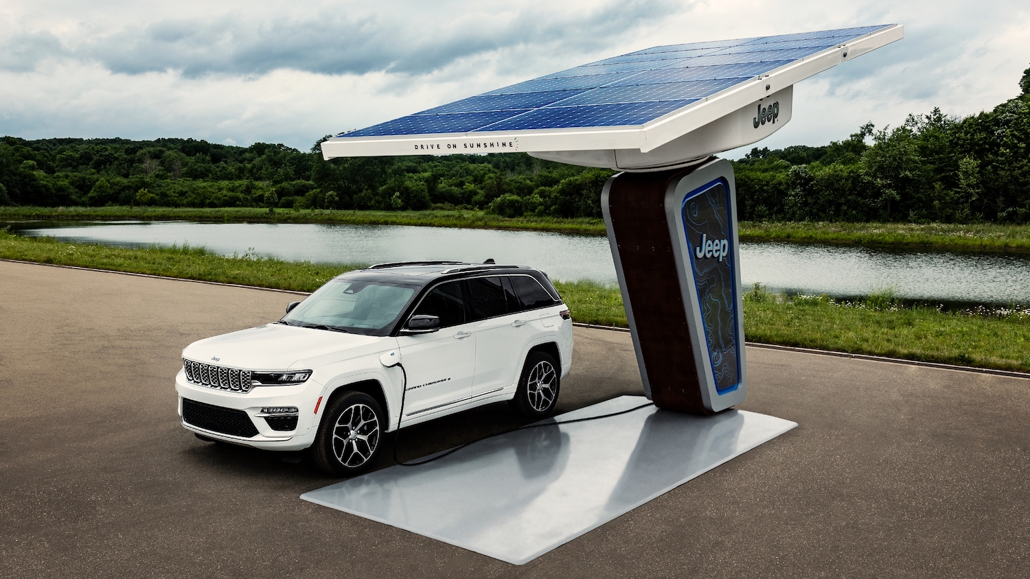 Jeep Grand Cherokee 4xe plug-in hybrid suv plugged into a solar charger by a pond, trees visible in the background.
