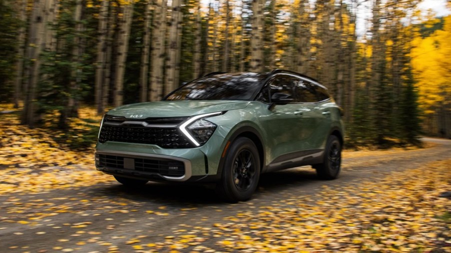 A green 2023 Kia Sportage small SUV is driving on a road surrounded by leaves.