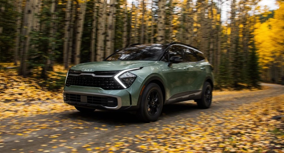 A green 2023 Kia Sportage small SUV is driving on a road surrounded by leaves. 