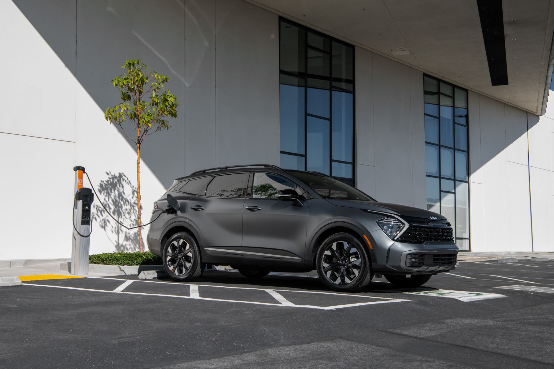 A dark gray 2023 Kia Sportage plug-in hybrid electric vehicle (PHEV) compact SUV plugged into a charging station