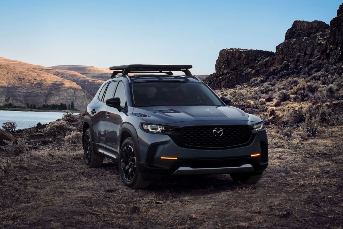 A 2023 Mazda CX-50 compact crossover SUV with roof rails parked on dirt near water and mountains