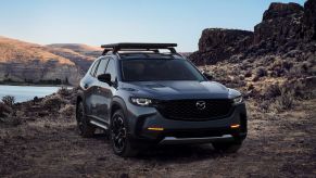 A 2023 Mazda CX-50 compact crossover SUV with roof rails parked in the dirt near water and mountains