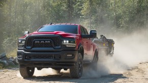 Red Ram 2500 Rebel heavy-duty off-road pickup truck towing a trailer of 4x4s through the woods, a dust cloud visible behind it.