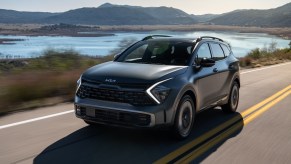 A grey 2023 Kia Sportage PHEV driving down a country road with mountains and water in the background.