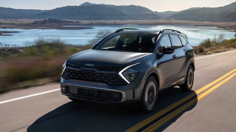 A grey 2023 Kia Sportage PHEV driving down a country road with mountains and water in the background.