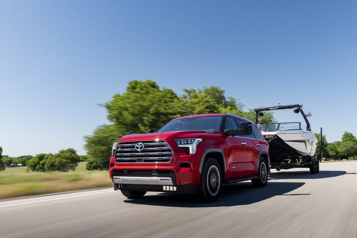 The red Toyota Sequoia SUV shows off its greater towing capacity than the entry-level Jeep Wagoneer Series I when pulling a boat down a country road.