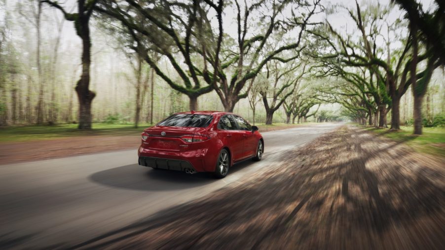 A rendering of a red 2023 Toyota Corolla Hybrid is driving away from the viewer along a treelined paved road.
