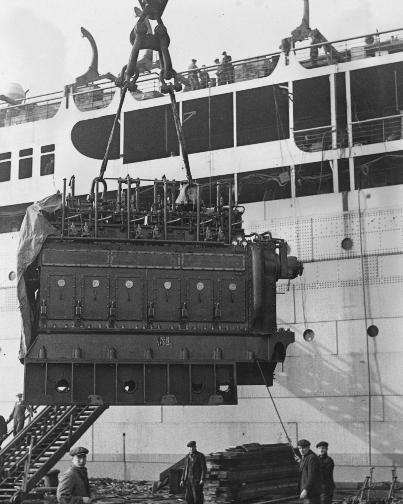 The world's first six-cylinder auxillary engine being lifted aboard the Athlone Castle ship by a crane, dockworkers looking on.