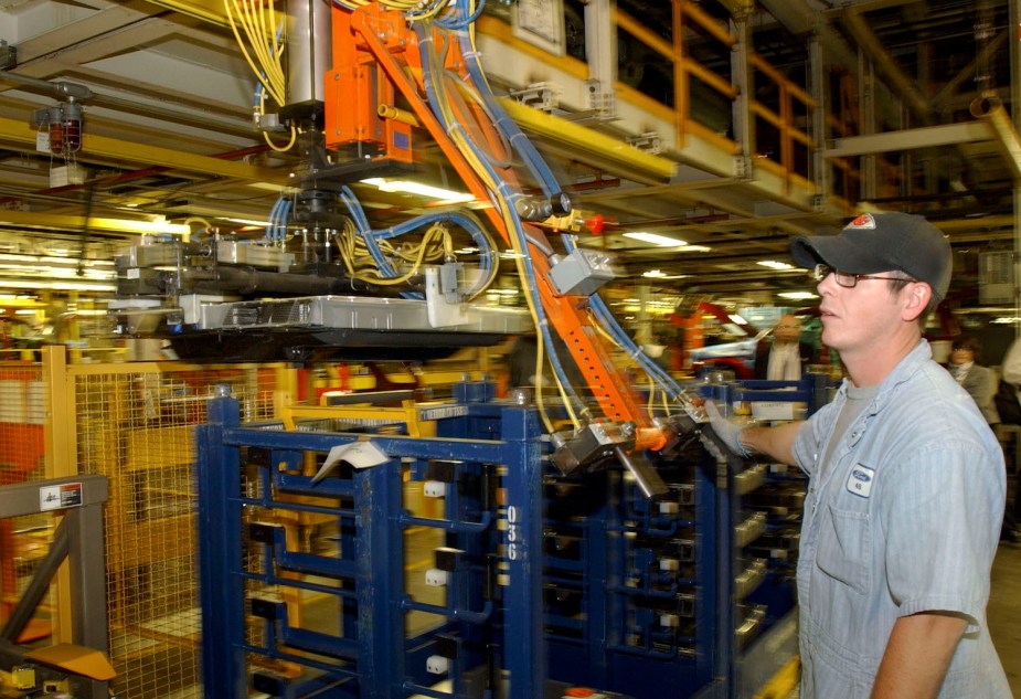 A Ford Motor Company factory worker uses a crane to move a Nickel Metal Hydride (NiMH) battery destined for a Mercury Mariner hybrid SUV, the blurry assembly line visible in the background.