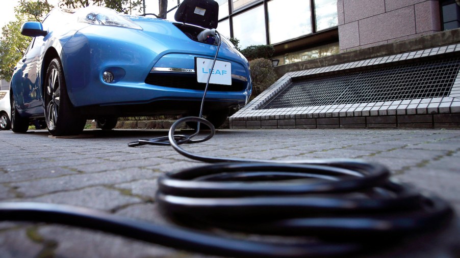 Promo photo of a blue 2013 Nissan Leaf charging in a driveway, a black charging cable coiled on the ground visible in front of the car.