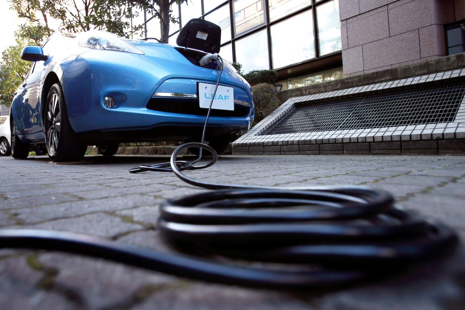 Promo photo of a blue 2013 Nissan Leaf charging in a driveway, a black charging cable coiled on the ground visible in front of the car.