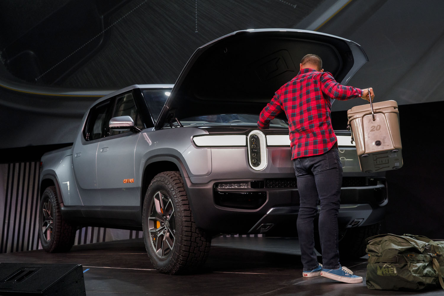 A man setting a cooler in the front trunk of a silver Rivian electric truck parked on a stage.