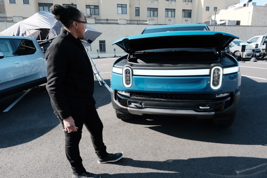 Woman holding a key fob and opening the frunk of a Blue Rivian R1T electric pickup truck in a parking lot.