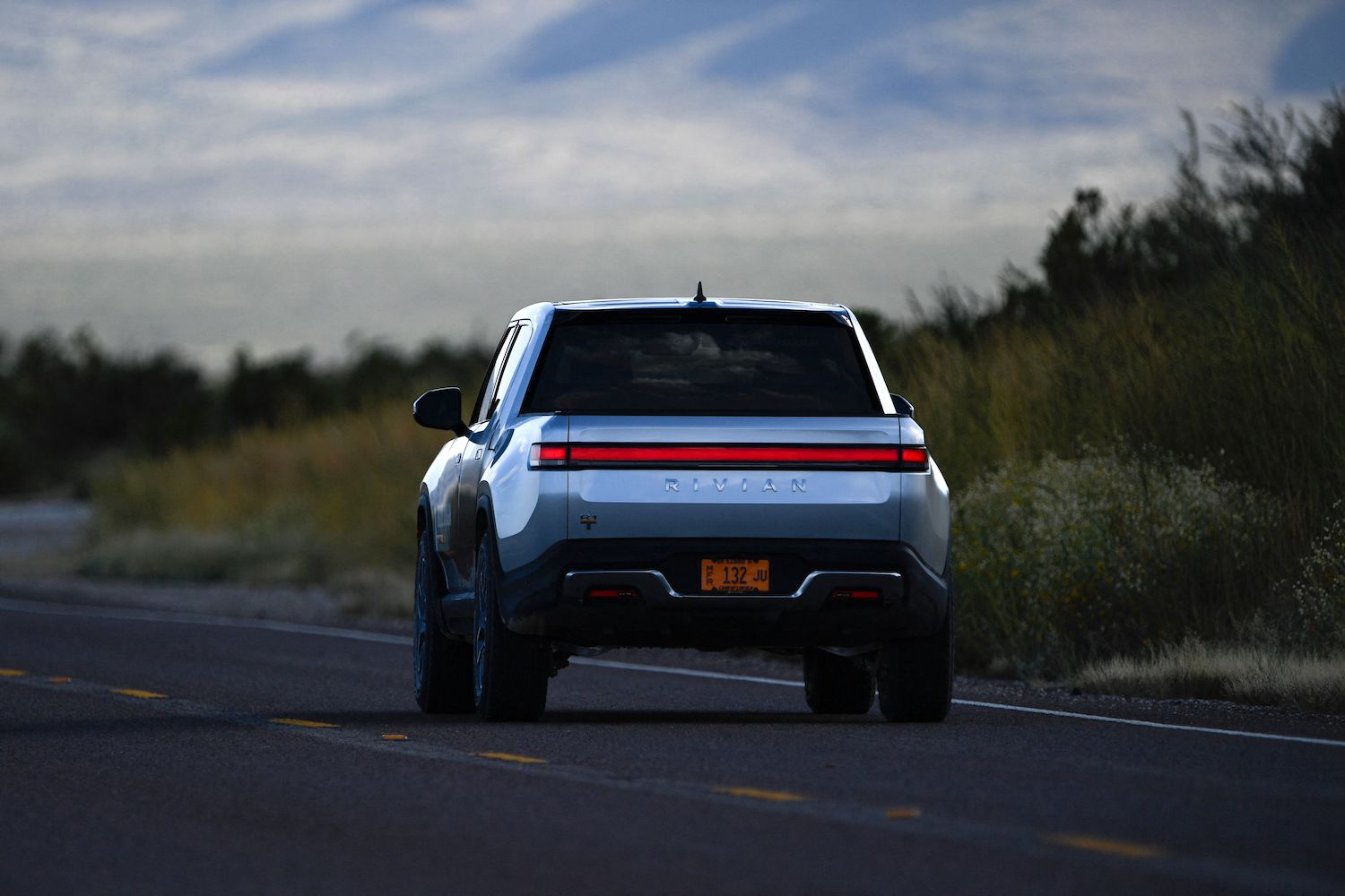 Silver Rivian truck driving away from the camera on a rural highway.