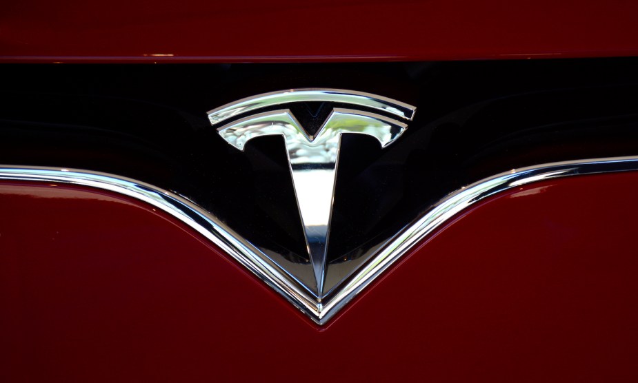 Closeup of the chrome "T" Tesla badge on a red, used electric vehicle.