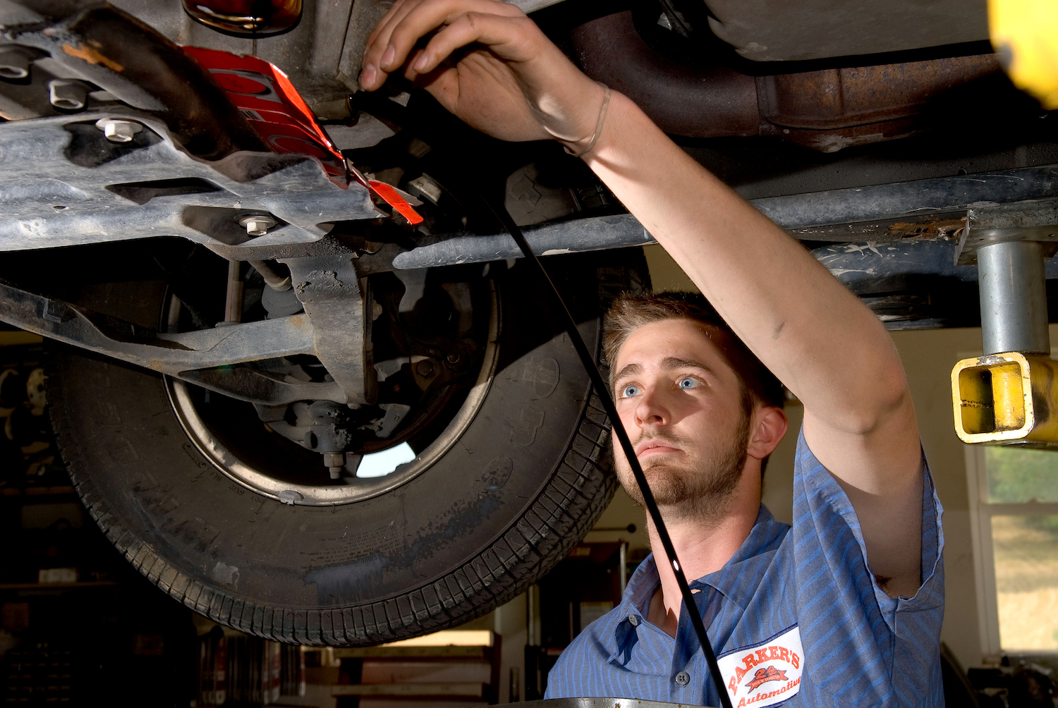 An auto technician maintains a vehicle on an overhead lift, one of its tires visible behind him.