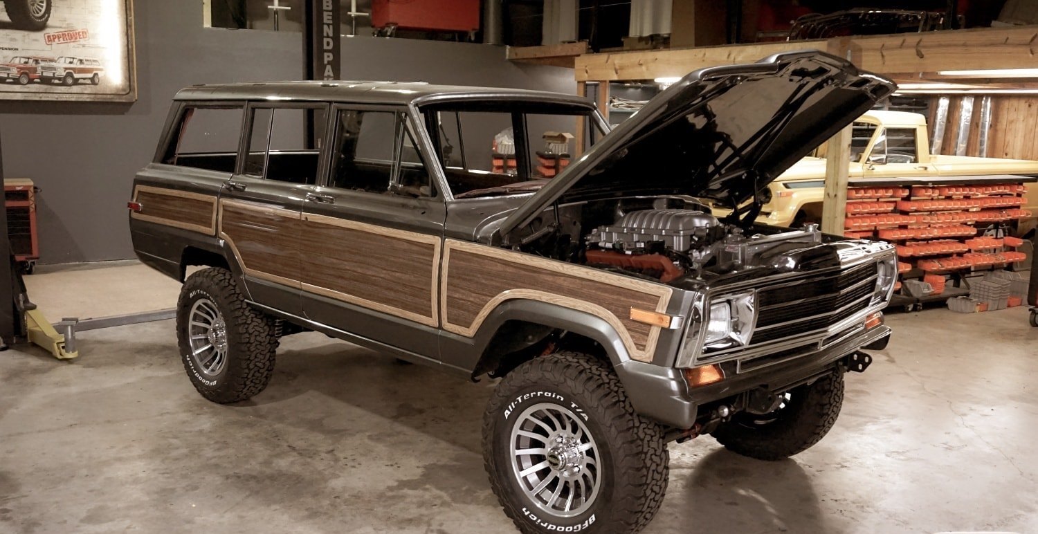 A Jeep Grand Cherokee restomod with a supercharged Hellcat V8 parked in a garage, a pickup truck and tools visible in the background.