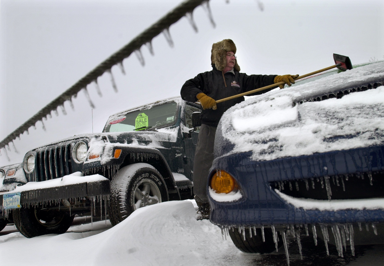 A car salesman brushes snow off of a car.